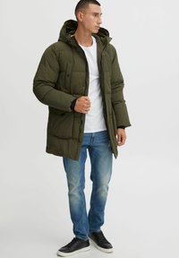 Chaqueta acolchada de color verde oliva con capucha, cierre de cremallera y bolsillos frontales, llevada sobre una camiseta blanca y jeans azules, con zapatillas negras.