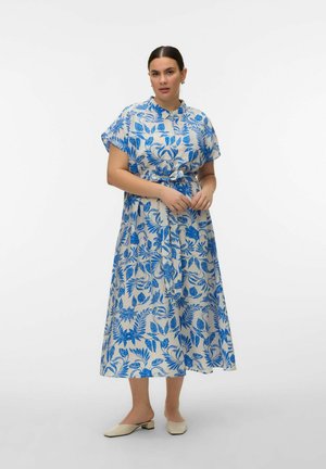 Frau steht und trägt ein weißes Midikleid mit blauem Blumenmuster, kurzen Ärmeln, gebundener Taille und beigen, flachen Slip-On-Schuhen.