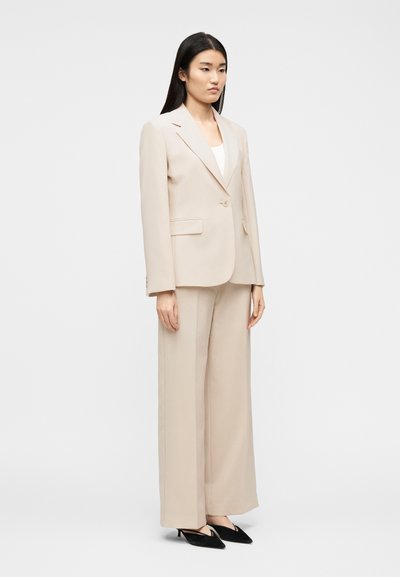 Γυναίκα που στέκεται, φοράει μπεζ προσαρμοσμένο σακάκι και παντελόνι με φαρδιά πόδια, με μαύρα χαμηλά παπούτσια μυτερά, σε απλό φωτεινό φόντο.