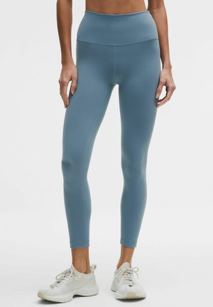 Legíny s vysokým pasem, bezšvové, přiléhavé, z měkké matné modré látky. Kombinovány se světle barevnými sportovními botami.