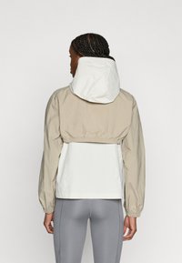 Ελαφρύ, δίχρωμο μπουφάν με μπεζ πάνω μέρος και λευκό κάτω μέρος, με κουκούλα και ελαστικά μανίκια για εφαρμοστή σιλουέτα.