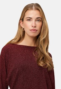 Haut en maille bordeaux avec un large col bateau, des manches longues, et une apparence texturée et scintillante, porté avec des boucles d'oreilles créoles dorées.