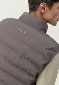 Bruine puffer vest met een hoge kraag, met verticale quilting en een reflecterende logo op de achterkant. De stof lijkt glad en mat.