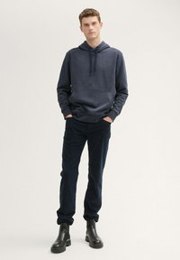 Felpa navy con tasca a marsupio e cappuccio regolabile, abbinata a pantaloni scuri. Modello che indossa stivali chelsea neri, in piedi su uno sfondo chiaro.