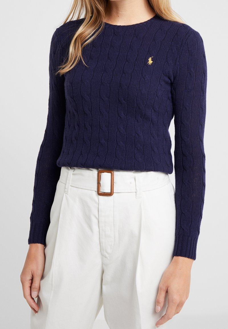 Femme portant un pull en maille torsadée bleu marine avec un logo brodé jaune, associé à un pantalon blanc taille haute et une boucle de ceinture marron.