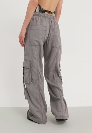 Persona con pantalones cargo grises sueltos con bolsillos traseros y laterales, combinados con una camiseta estampada sin espalda y calzado oscuro.