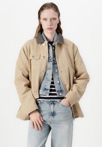 Béžová plátěná bunda s šedým kordovým límcem, vrstvená přes světle modrou džínovou vestu. Pod ní pruhovaná košile, světle modré džínové kalhoty, neutrální pozadí.