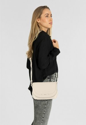 Femme aux longs cheveux blonds portant un chemisier noir et un jean gris, avec un petit sac beige porté en bandoulière.