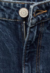 Gros plan sur la ceinture d'un jean en denim bleu avec un bouton en métal marqué "MNGDNW 1984" et une fermeture éclair partiellement ouverte.