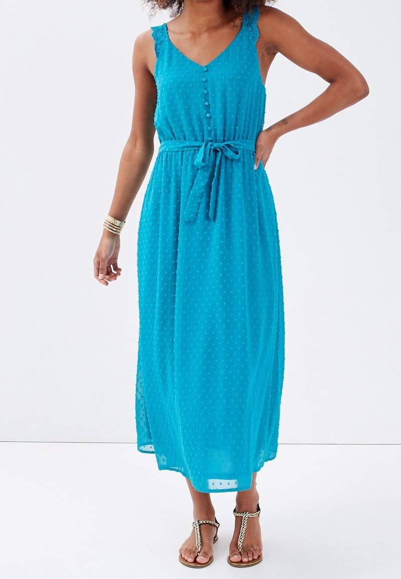 Femme portant une robe midi turquoise sans manches avec des boutons et une ceinture à la taille, associée à des sandales dorées tressées et des bracelets.