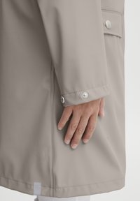 Beige vanntett frakk med en glatt tekstur, som har en knappet mansjett og en lomme. Stoffet er litt blankt med en rett kutting.