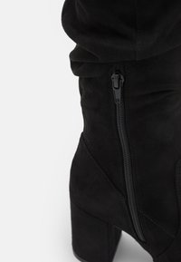 Svart mocka ankelsko med en staplad klack, som har en sidodragkedja och en mjuk, texturerad yta. Designad med subtil rynkning upptill.