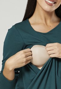 Teal langærmet top med bådudskæring, trukket til side for at afsløre en beige bh, der fremhæver stoffets tekstur og pasform.