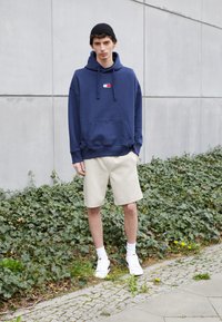Felpa navy con tasca frontale e cordini, pantaloni corti beige, sneakers bianche e berretto nero, su uno sfondo di edera verde.