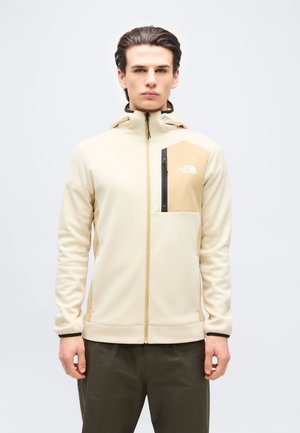 Joven con sudadera beige con cremallera y bolsillo en el pecho con el logo de The North Face, posando frente a un fondo claro y liso.