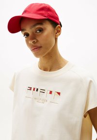Czerwona bawełniana czapka z zakrzywionym daszkiem. Model ma na sobie jasną, kremową koszulkę z haftowanymi flagami i złotym napisem "HILFIGER".