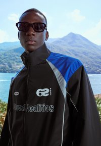 Czarna kurtka z niebieskimi i szarymi akcentami, z wysokim kołnierzem, z zamkiem błyskawicznym z przodu oraz tekstem "Altered Realities" i "62". Duże czarne okulary przeciwsłoneczne.