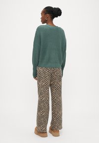Πράσινο πλεκτό πουλόβερ με άνετη εφαρμογή, συνδυασμένο με παντελόνια φαρδιάς γραμμής με καφέ και μαύρο ζιγκ-ζαγκ σχέδιο. Λείο υφασμάτινο υλικό.