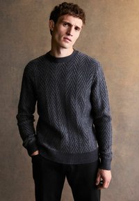 Dunkelgrauer Pullover mit Fischgrätmuster, Rundhalsausschnitt, gerippte Bündchen und Saum, aus strukturiertem Strickstoff, getragen mit schwarzen Hosen.