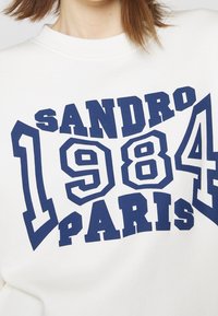 Kermansweater med djärv marinblå text som läser "SANDRO PARIS 1984" i en geometrisk design. Mjuk, slät textur, klassisk rund halsringning.