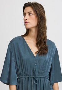 Blaues V-Ausschnittkleid mit kurzen, weiten Ärmeln und einer Kordelzugtaille. Hergestellt aus einem glatten, glänzenden Stoff mit einer fließenden Silhouette und subtiler Textur.