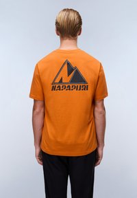 Orange bomulds t-shirt med et stort sort bjerg logo og "NAPAPIURI" tekst på ryggen. Korte ærmer, rund hals, afslappet pasform.