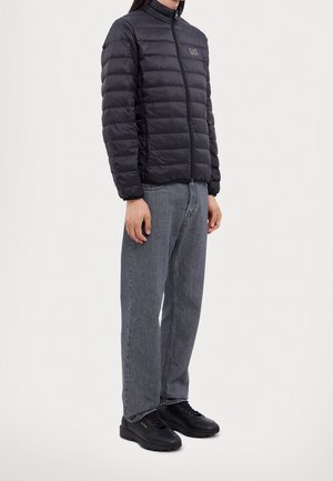 Personne portant une veste noire matelassée avec une fermeture éclair, un jean droit gris et des baskets noires, debout devant un fond clair uni.