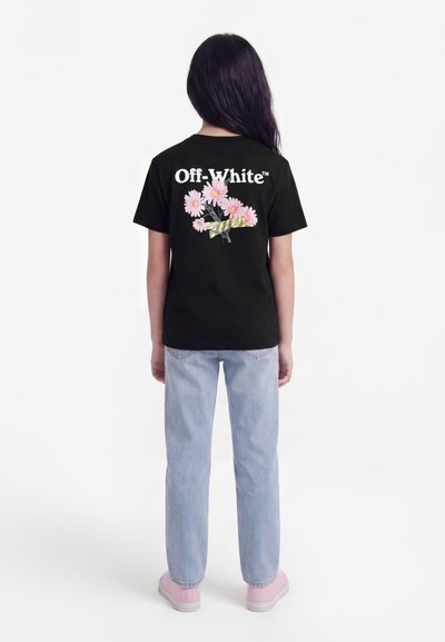 Άτομο με μακριά σκούρα μαλλιά φοράει μαύρο T-shirt Off-White με ροζ λουλούδια και σχέδιο με ταινία στο πίσω μέρος, ανοιχτό μπλε τζιν και ροζ παπούτσια.