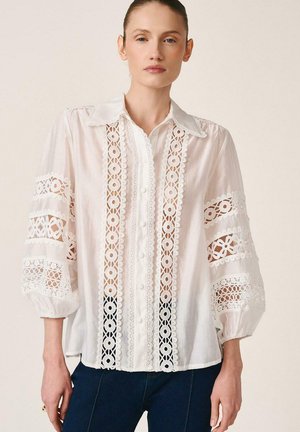 Mujer vestida con una blusa blanca con paneles de encaje en la parte delantera y en las mangas abullonadas tres cuartos, combinada con pantalones azul oscuro.