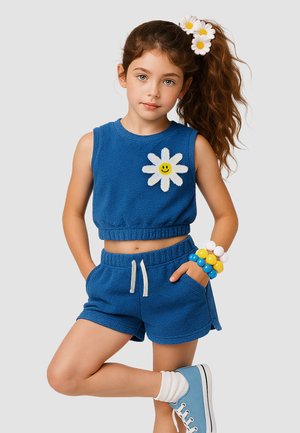 Junges Mädchen mit langen Haaren, die mit Gänseblümchen-Haarklammern gebunden sind, trägt ein blaues ärmelloses Top und Shorts mit einem lächelnden Gänseblümchen-Motiv und bunte Perlenarmbänder.