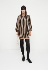 Kvinde iført en kjole med beige og sort zigzag-mønster, sorte strømpebukser, sorte spidse hæle, som holder en lille hvid taske, stående foran en hvid baggrund.