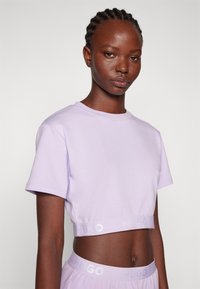 T-shirt cropped lavande en coton doux avec des manches courtes, arborant un ourlet élastique avec logo. Texture lisse et coupe décontractée.