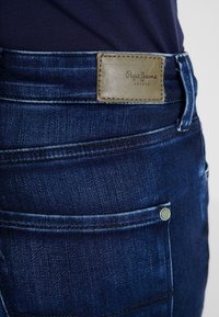 Tmavě modré džínové kalhoty s texturovanou tkaninou, s hnědou koženou visačkou s nápisem "Pepe Jeans London" na pase.