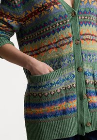 Stickad kofta i grönt med flerfärgade geometriska mönster, ribbad fåll och muddar, fem träknappar och sidfickor.
