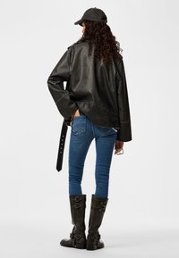 Femme aux longs cheveux bouclés portant une veste en cuir noire, un jean skinny bleu, des bottes noires à boucle et une casquette noire, debout avec une main dans la poche.