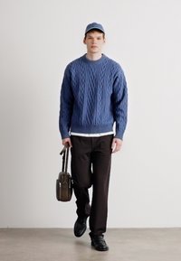 Blauwe kabelgebreide trui, witte shirt eronder, zwarte broek. Bruine tas in de hand, draagt een blauwe pet en zwarte schoenen. Eenvoudig, modern ontwerp.
