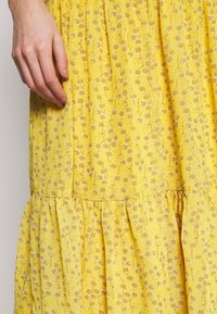 Kvinde hånd hviler på gul kjole med små hvide blomstermønstre og indsamlet sømdetalje.