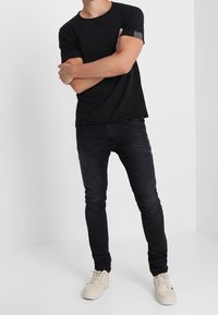 Sort kortærmet t-shirt med en tekstureret lap på venstre ærme, parret med sorte slim-fit jeans og lyse beige sneakers.