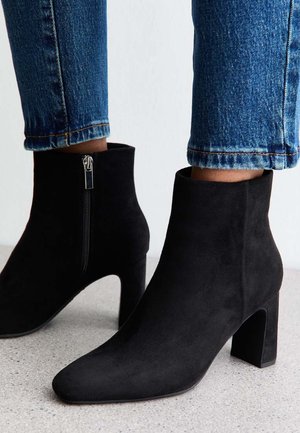 Stivaletti neri in suede con punta appuntita e tacco spesso. Presentano una cerniera laterale e una superficie testurizzata, abbinati a jeans blu aderenti.