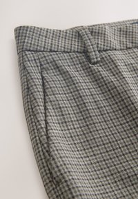 Gros plan sur un pantalon en tissu à carreaux gris et noir avec une ceinture, des passants et une poche latérale sur un fond neutre.