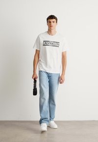 Young man walks forward holding a black belt bag, wearing a white Calvin Klein t-shirt, light blue jeans, and white sneakers.