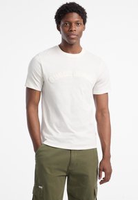 Weißes Baumwoll-T-Shirt mit leichtem gesticktem "Guess Jeans"-Logo auf der Brust. Kurze Ärmel und runder Ausschnitt, getragen mit grünen Cargo-Shorts.