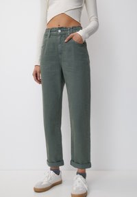Pantalon en denim vert avec taille élastique, revers retroussés, poches avant et fermeture à bouton. Porté avec des baskets blanches.