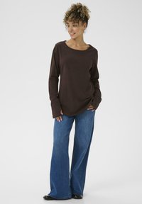 Langærmet brun bluse med rund halsudskæring, parret med blå flare jeans. Stoffet ser blødt og glat ud, hvilket fremhæver en afslappet pasform.