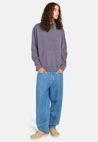 Lila Baumwollkapuzenpullover mit Kängurutasche, kombiniert mit lässigen hellblauen Denim-Jeans und beigen Slipschuhen.