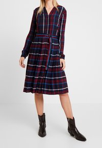 Kobieta w długiej koszulowej sukience w kratę z paskiem w talii i ciemnobrązowych botkach za kostkę, stojąca na tle białym.