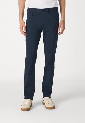 Pantalones de algodón azul marino con un corte slim, cinco bolsillos y cierre de botón. Acompañados de zapatillas de color crema con suelas de goma.