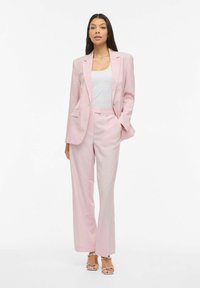Veste et pantalon sur mesure rose pâle, avec un haut blanc. Le tissu a une texture lisse et le costume présente des détails discrets au niveau des poches.