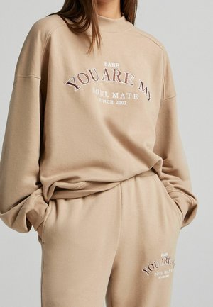 Femme portant un sweat-shirt et un pantalon beige avec le texte « Babe You Are My Soul Mate Since 2001 » imprimé sur les deux vêtements.