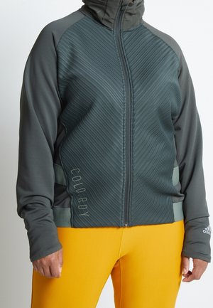 Persoon draagt een donkergrijs doorgestikt jack met rits en de tekst "COLD RDY" aan de zijkant, en gele legging, staat met ontspannen armen.